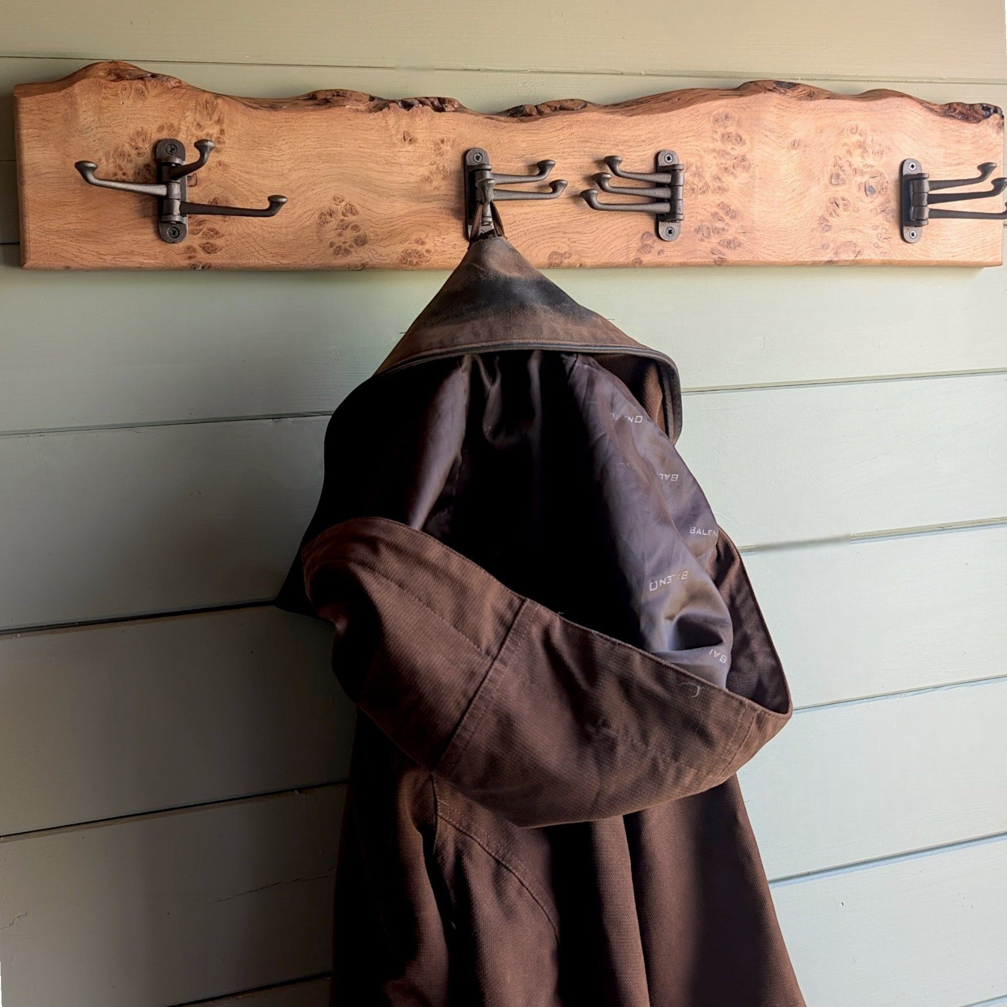 Thriverton hand-crafted Pippy Oak coat rack with live-edge profile and cast-iron hooks holding a brown hooded jacket.